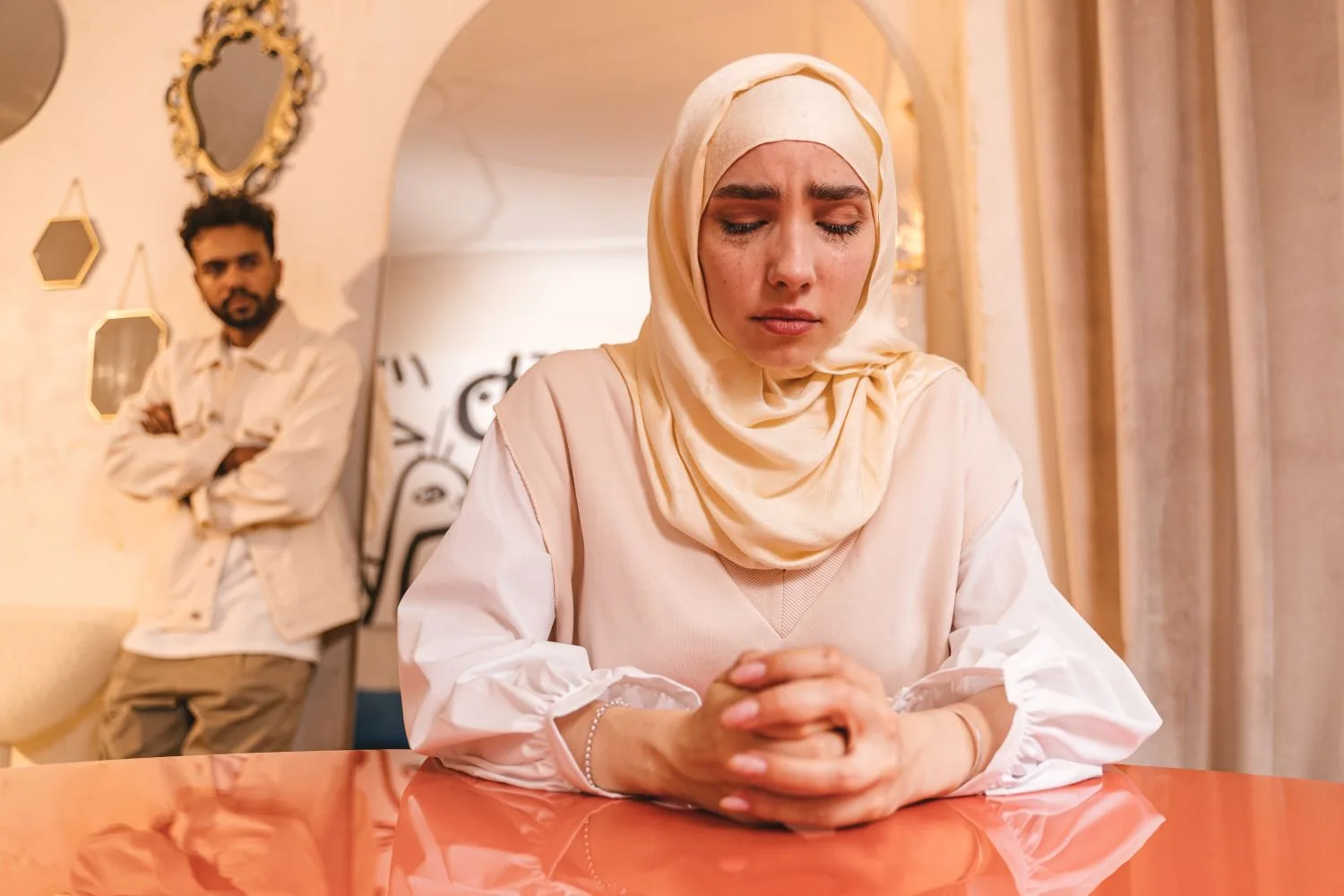 Une femme en hijab assise à une table, les mains jointes, le regard baissé ; derrière elle, son mari se tient debout, les bras croisés, à distance.
