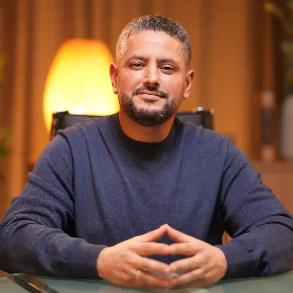 Abdessalam Hassani, assis à une table, les mains jointes, dans une pièce boisée éclairée par une lampe.