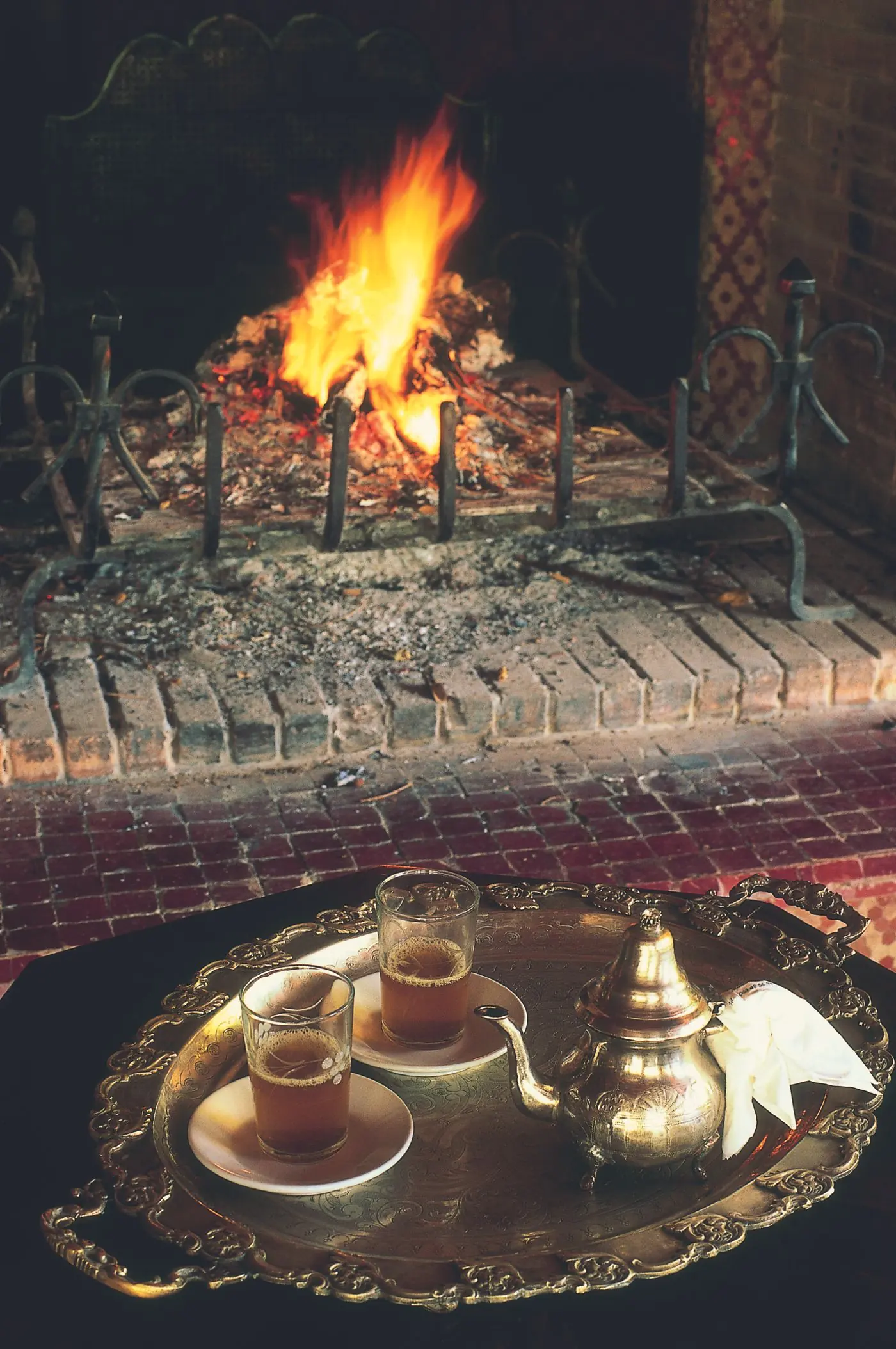 Deux verres de thé sur un plateau en laiton, posés devant un feu de cheminée.